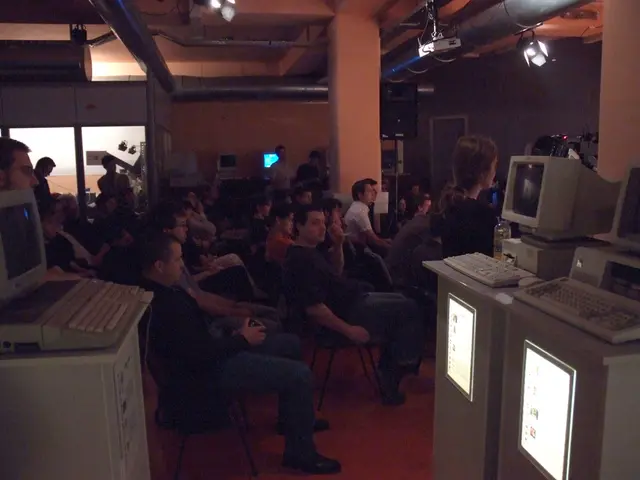 In the image there are many people sitting in a room and around them there are computers, speaker,...